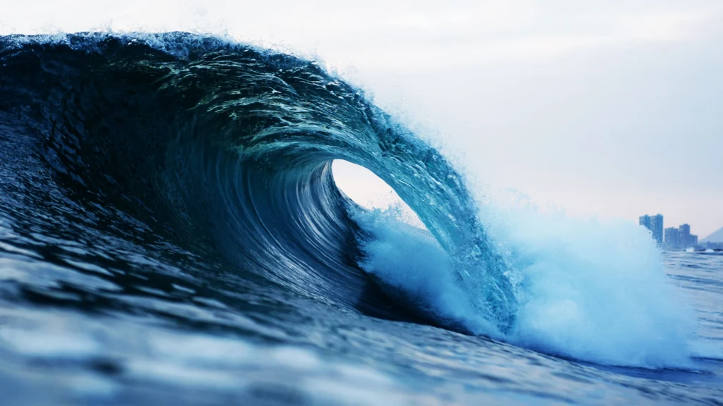 A large ocean wave breaking, with a city skyline faint on the horizon